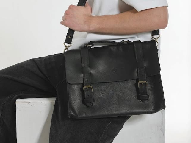 Model seated on a white block holding a Kankia black leather satchel briefcase with brass buckles