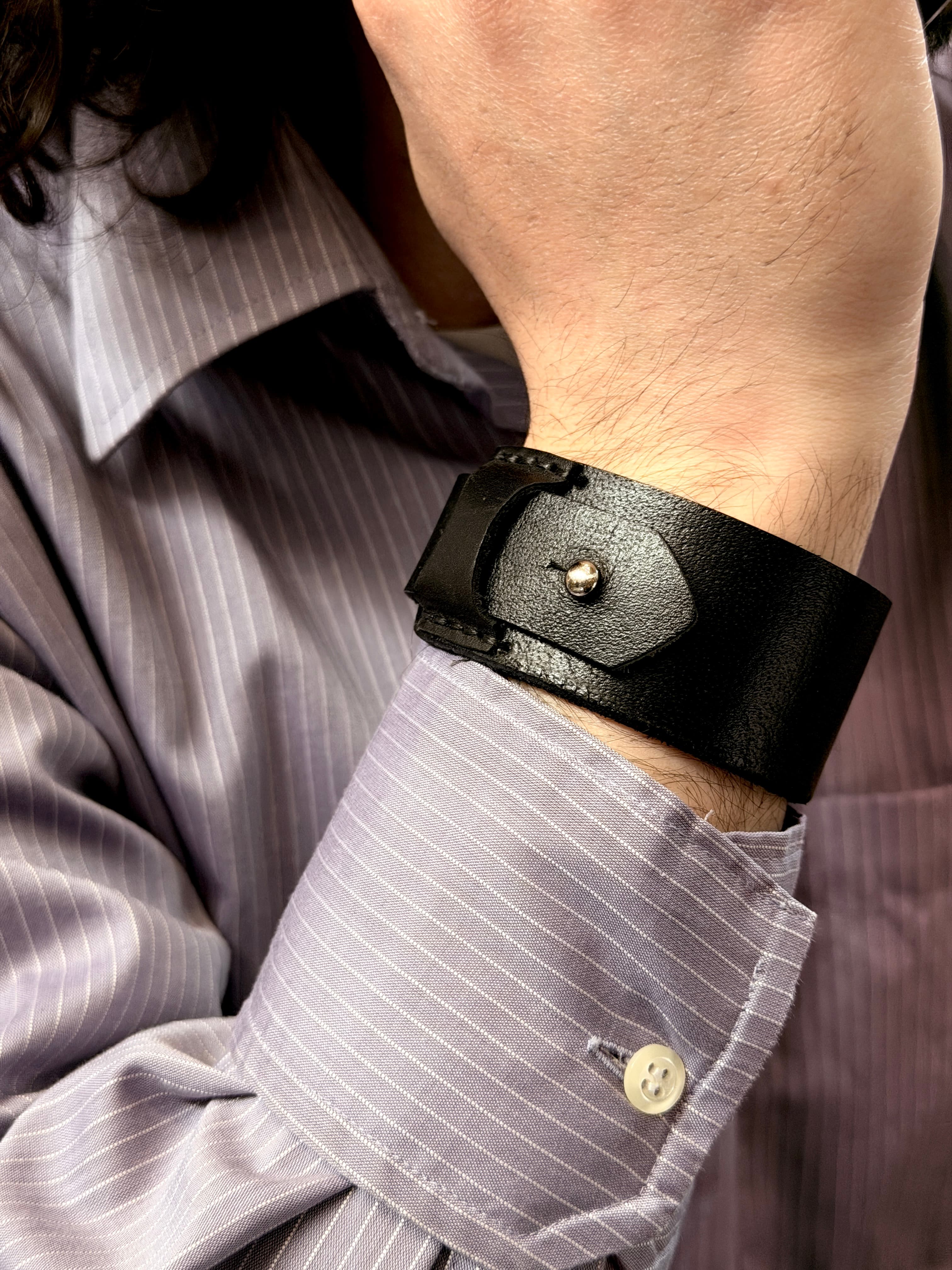 Close-up of a Kankia black leather cuff bracelet worn on a wrist, pinstripe shirt detail